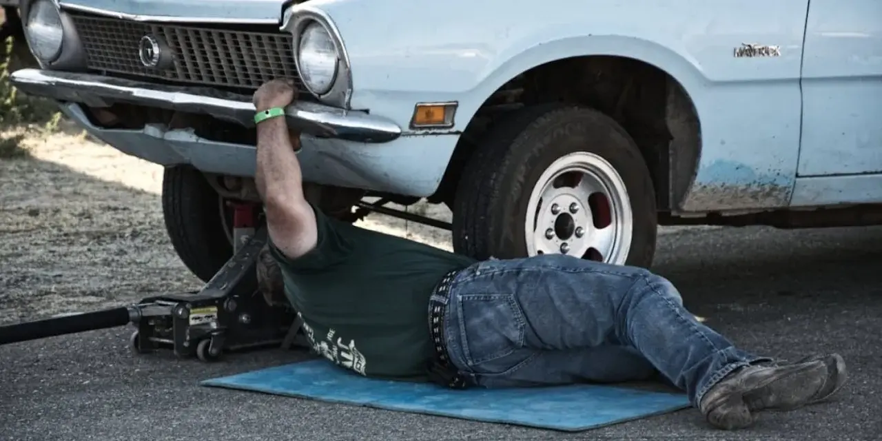 Un uomo lavora sotto un'auto d'epoca, forse per un intervento di **art 46 C.d.S.**, con un cric che la solleva.