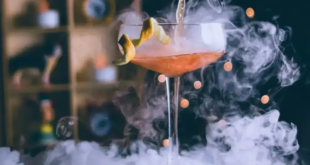 Kieliszek z drinkiem otoczony gęstym dymem do drinków, z pomarańczową skórką jako dekoracją.