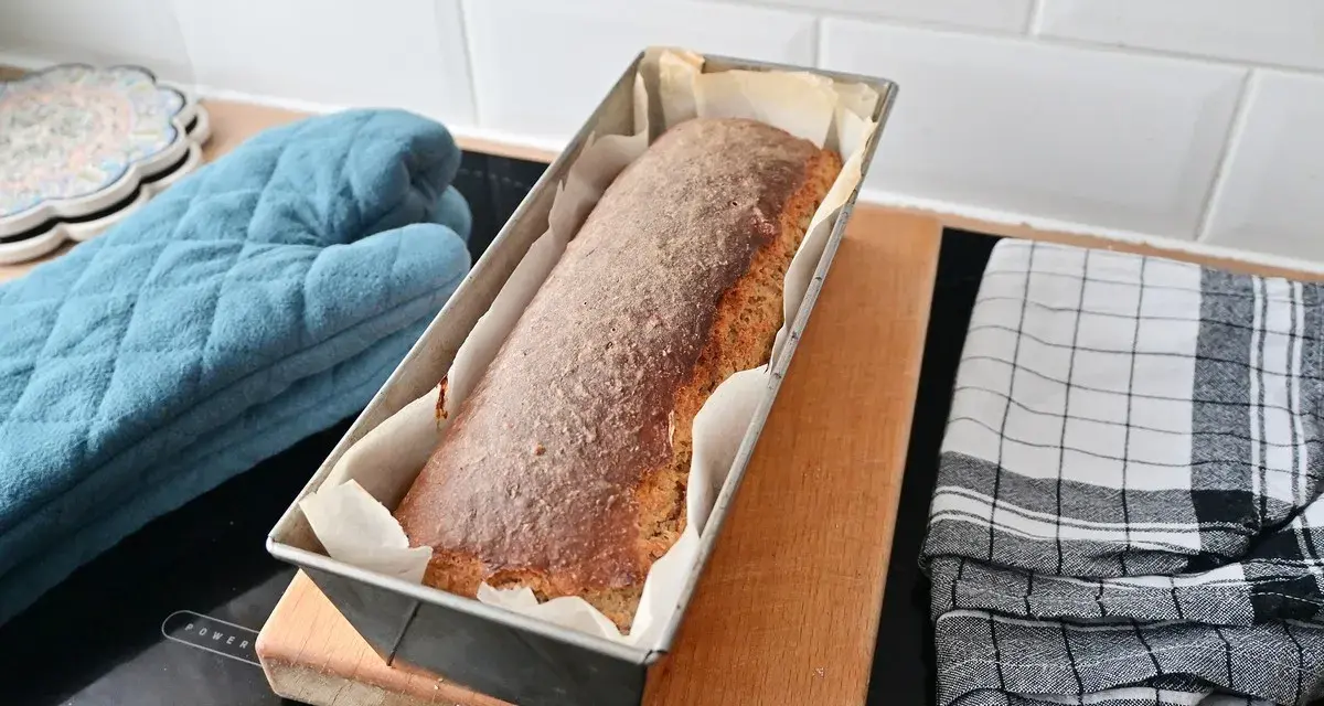Świeżo upieczony chleb cebulowy na zakwasie w keksówce, gotowy do wyjęcia. Obok rękawica kuchenna i ściereczka.