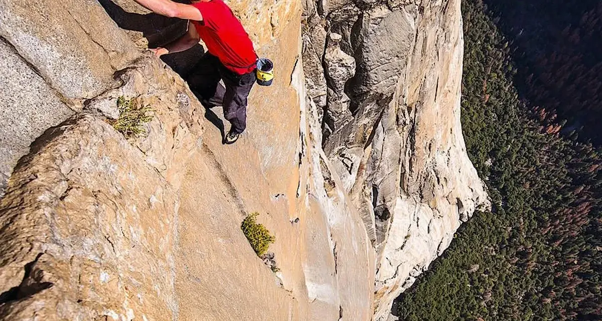 Ein Mann klettert free solo eine steile Felswand empor, mit einem tiefen Abgrund unter ihm.