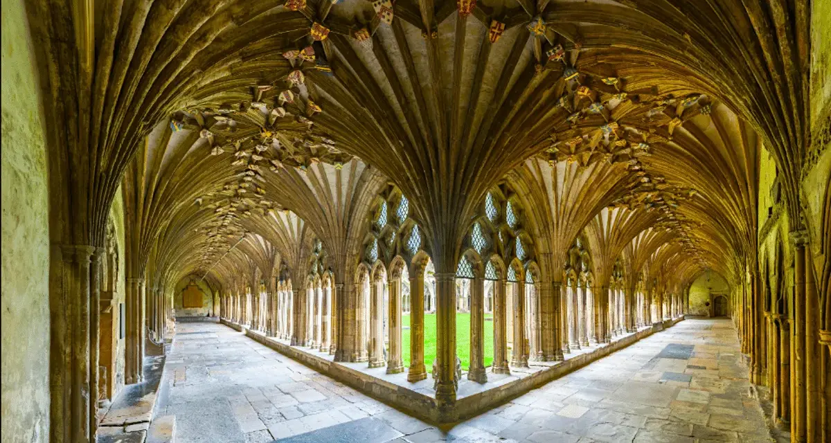 A grand cloister with vaulted ceilings, reminiscent of a monastery floor plan. Sunlight streams through arched windows onto the stone floor.