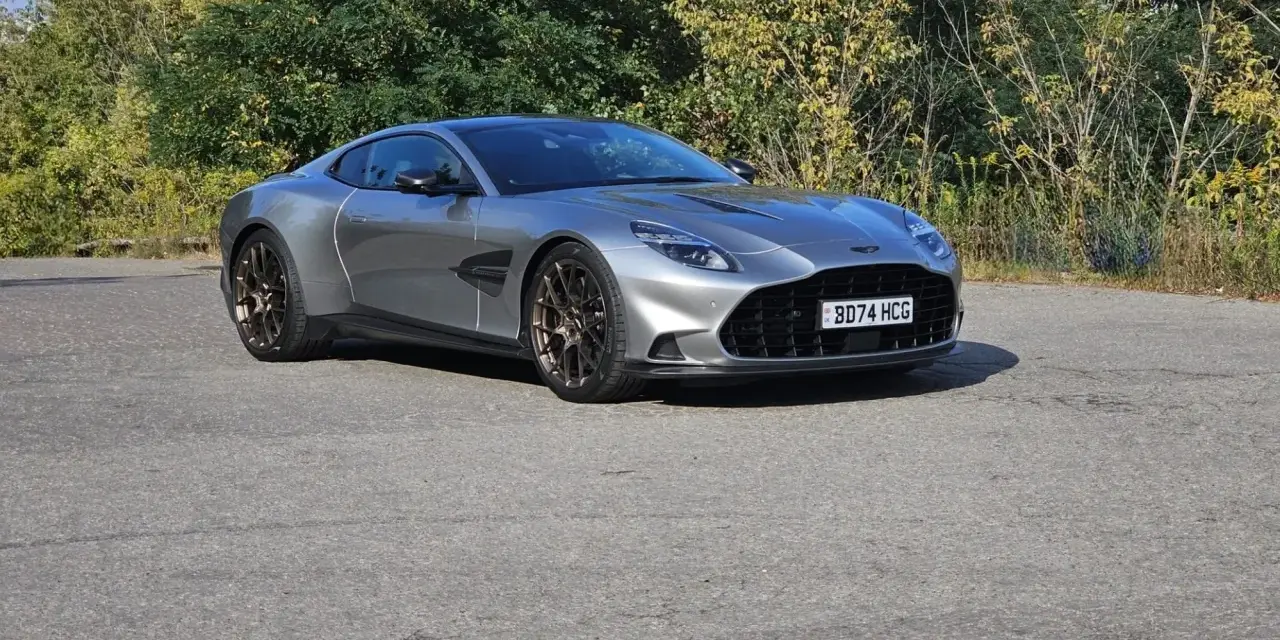 Srebrny Aston Martin Vanquish 2025 z brązowymi felgami na tle zieleni.