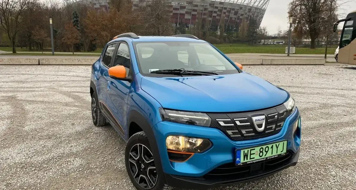 Niebieski Dacia Spring z pomarańczowymi akcentami na tle Stadionu Narodowego. Sprawdź atrakcyjną dacia spring cena!