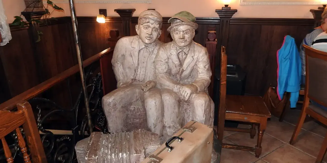 Dwie rzeźby siedzących mężczyzn, przypominających tońcio lub szczepcio, z akordeonem i walizką u stóp.