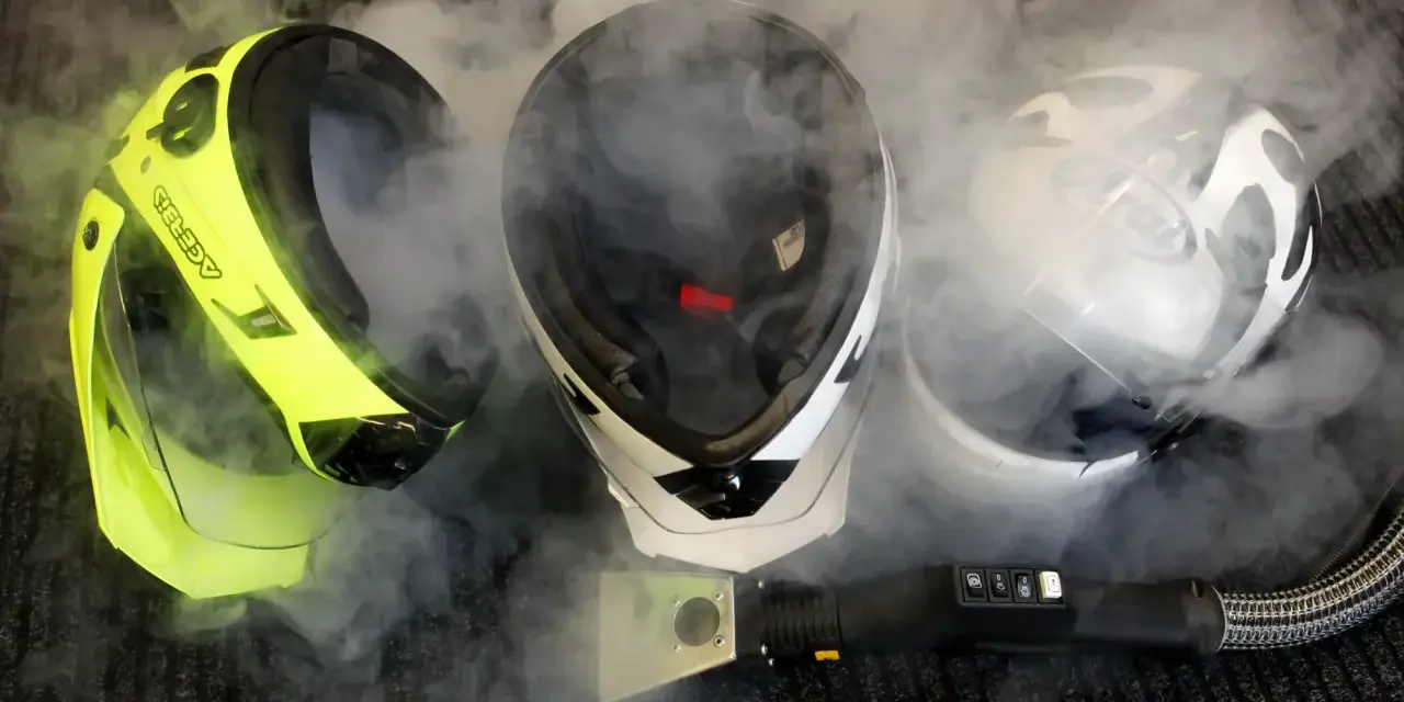 Pulizia del casco con vapore. Tre caschi, uno giallo fluo, uno bianco e nero, uno bianco e nero con visiera, circondati da fumo.