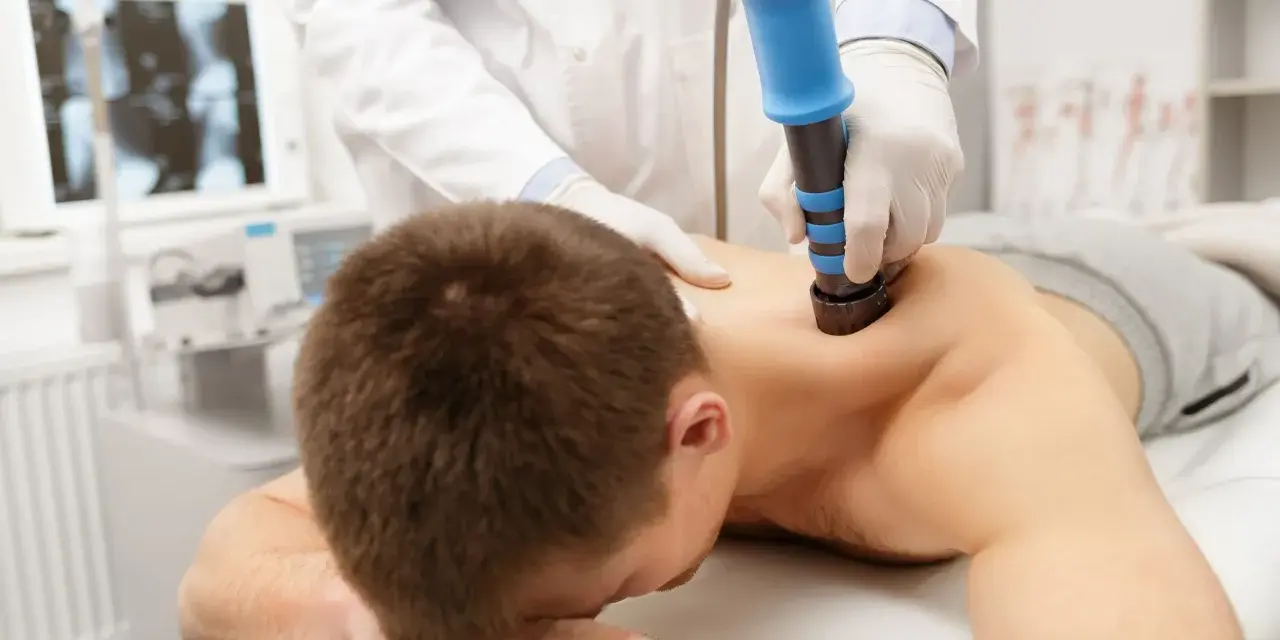 Arzt behandelt HWS-Syndrom mit Stoßwellentherapie. Ein Mann liegt auf dem Bauch, während ein Arzt mit einem blauen Gerät auf seinen Rücken drückt.