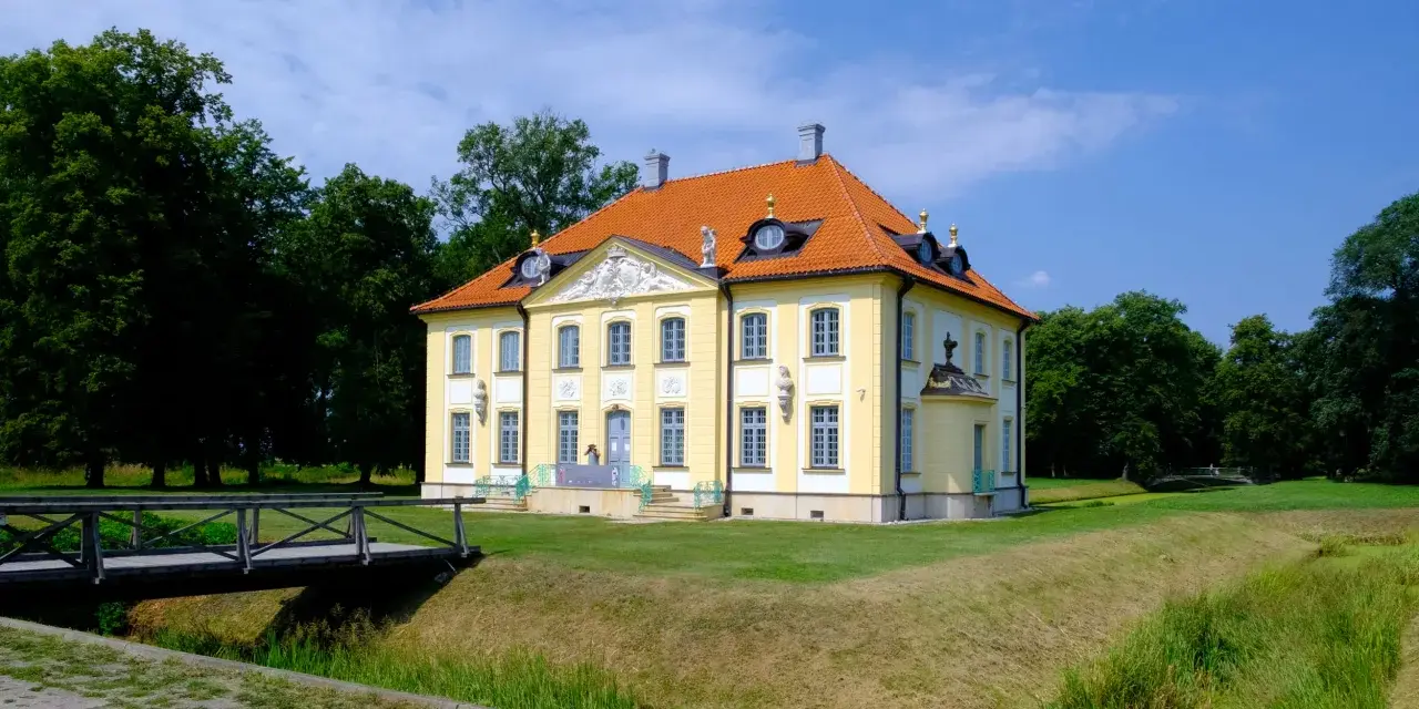 Pałac w Choroszczy, żółty, z pomarańczowym dachem, otoczony zielenią. Drewniany mostek prowadzi do posiadłości.