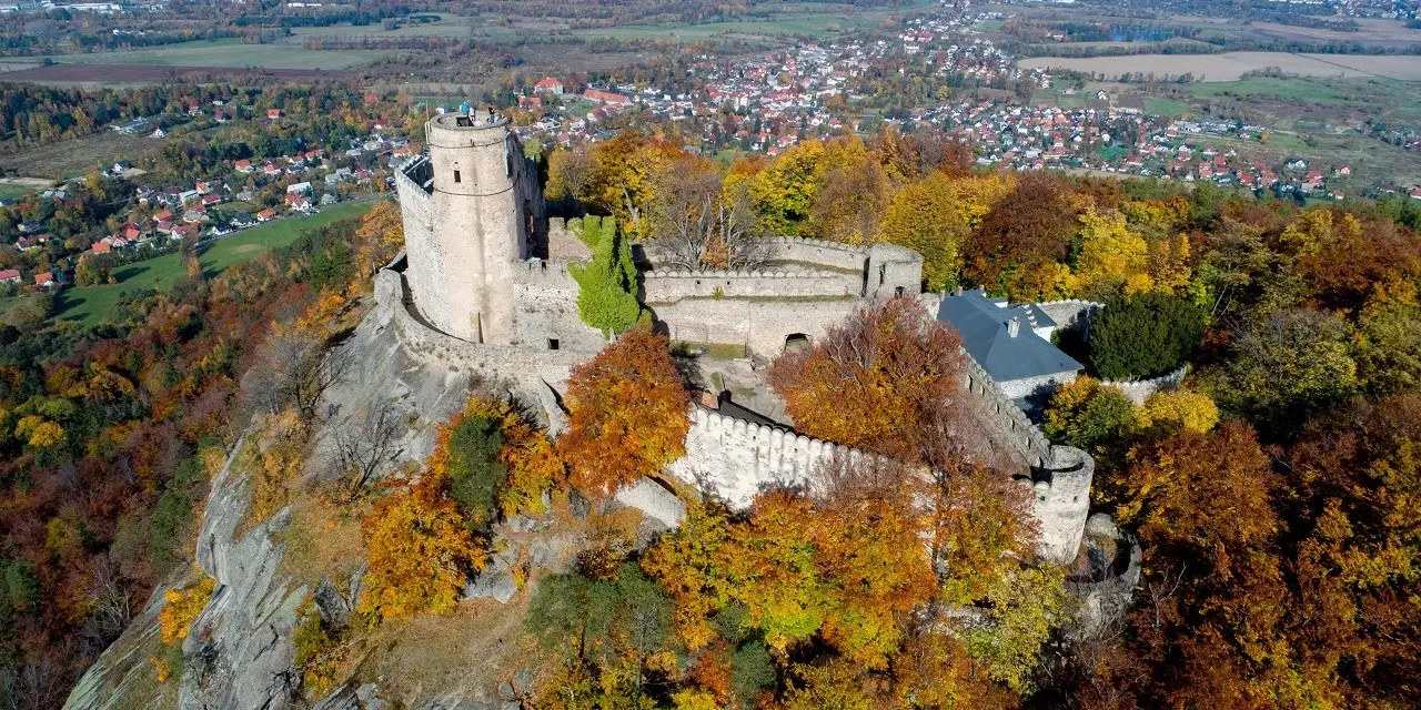 Zamek Chojnik na skalistym wzgórzu, otoczony jesiennymi barwami drzew. W oddali widać miasteczko i pola.