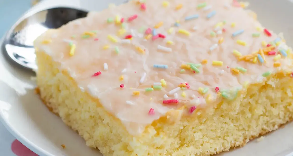 Ein Stück saftiger Fantakuchen vom Blech, mit Zuckerguss und bunten Streuseln bestreut, auf einem Teller mit Löffel.