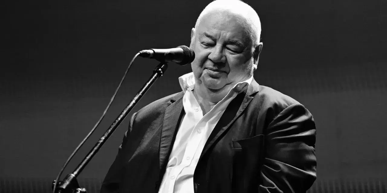 Czarno-białe zdjęcie Stanisława Sojki przy mikrofonie. Artysta z zamkniętymi oczami, w białej koszuli i marynarce, śpiewa. Stanisław Sojka nie żyje.