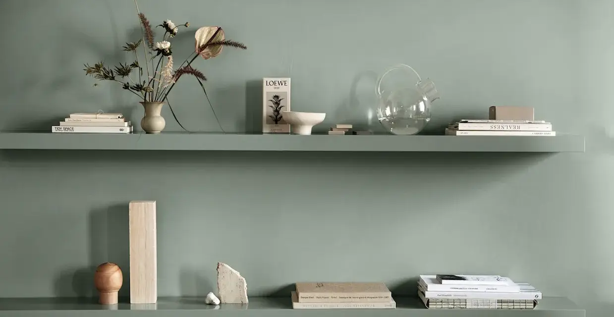 Två gröna hyllplan med böcker, vas med blommor, glasburk och dekorativa föremål. Enkel och stilren inredning.