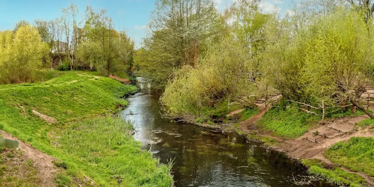 Ein idyllischer Panke Wanderweg schlängelt sich durch üppiges Grün. Ein kleiner Fluss fließt friedlich, gesäumt von Bäumen und Wiesen.