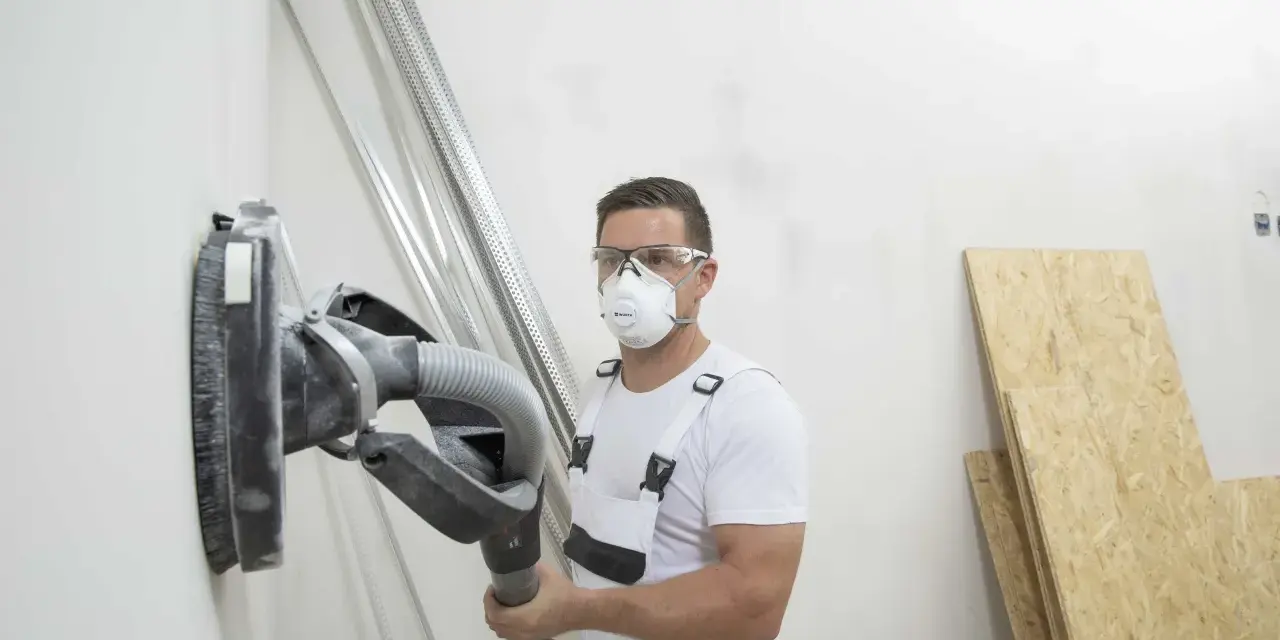 Operaio con maschera e occhiali sta per scartavetrare muro con una levigatrice.