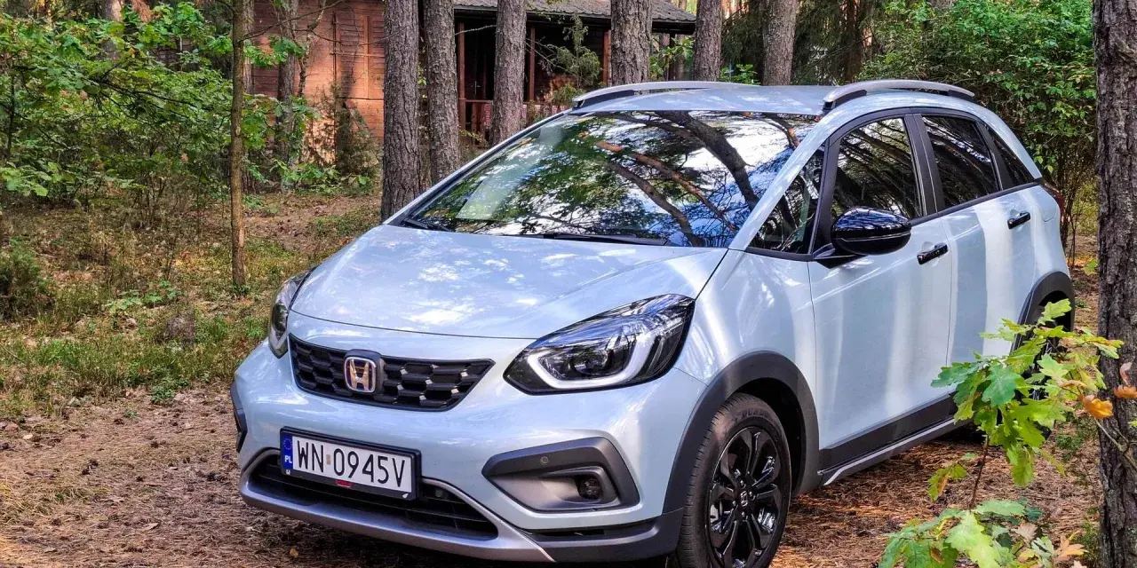 Jasny Honda Jazz Crosstar zaparkowany w lesie, obok drewnianego domku.