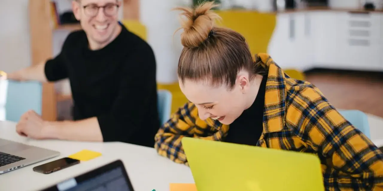 Zwei lachende Kollegen am Schreibtisch mit Laptops. Ein bezahltes Auslandspraktikum kann so Spaß machen!