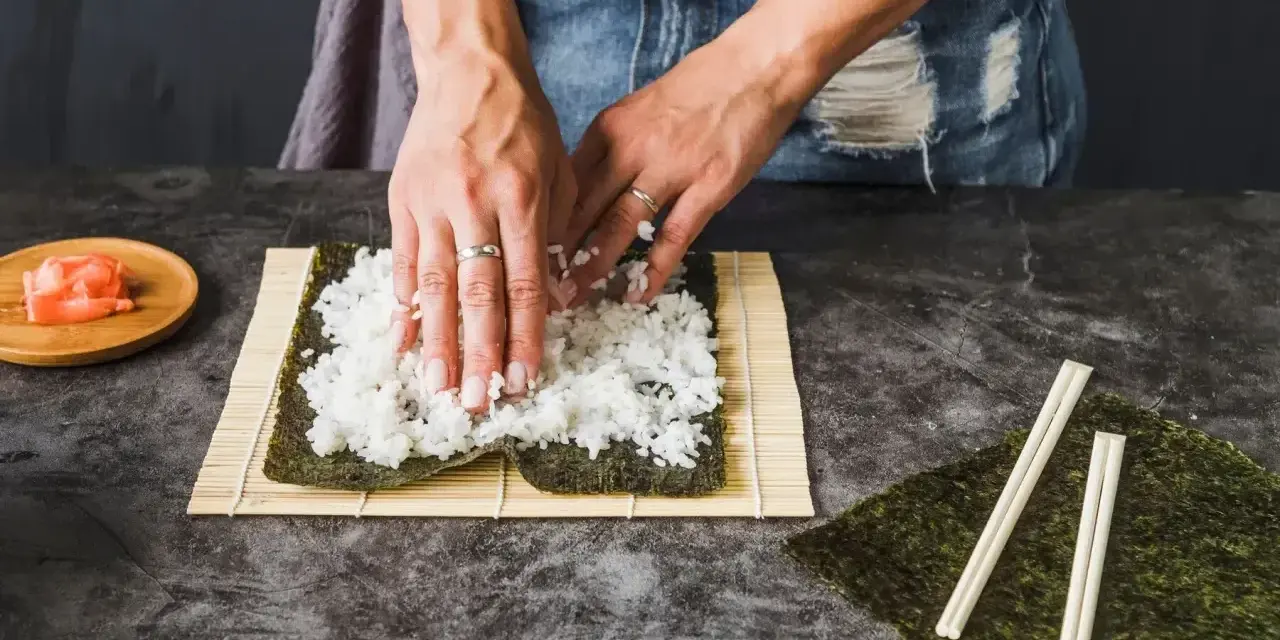 Ręce rozkładają ryż na nori, przygotowując się do zwinięcia sushi. Obok leżą pałeczki i talerzyk z imbirem, idealny dodatek do sushi.