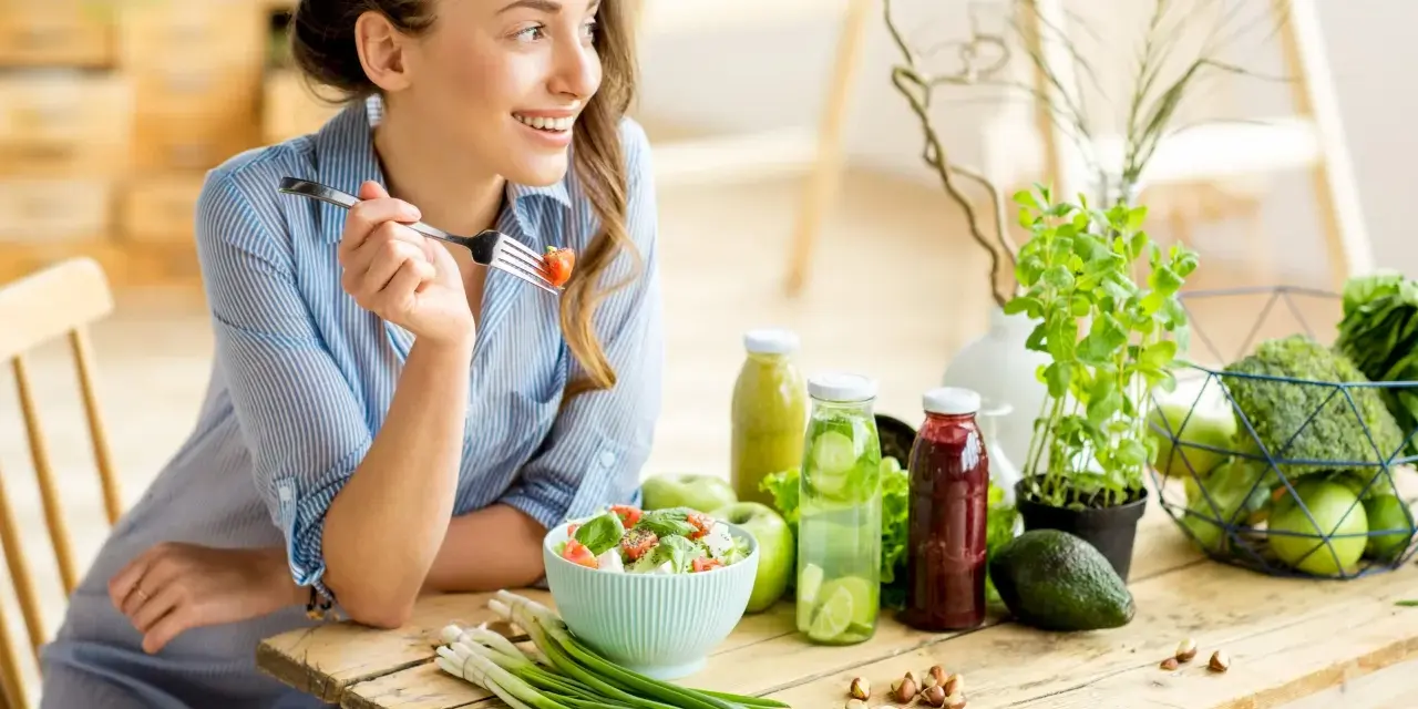 Kvinna äter sallad, omgiven av hälsosam mat. Kan vara en del av en kost för att undvika brist på kisel symtom.