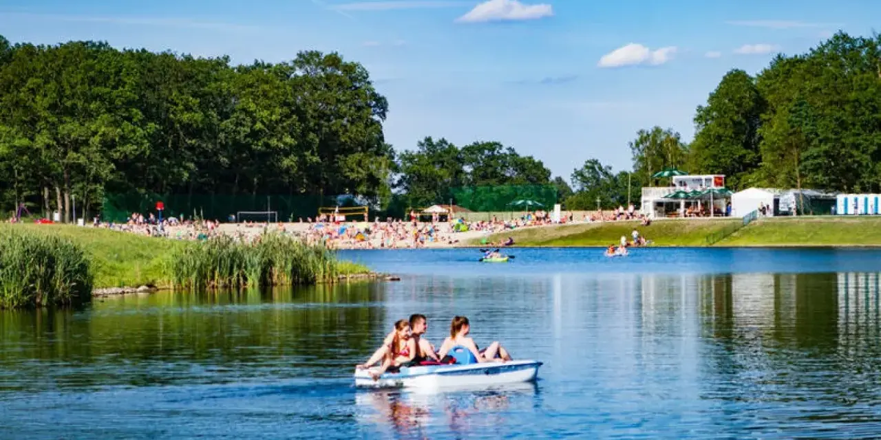 Rodzina na rowerku wodnym na jeziorze, plaża pełna ludzi, las i budynki w tle. Idealne miejsce gdzie nad wodę Dolny Śląsk.