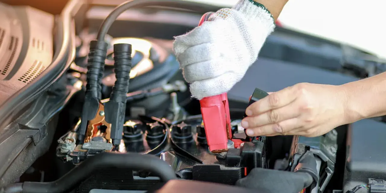 Eine Hand mit Handschuh klemmt das rote Kabel an den Pluspol, während die andere Hand den Minuspol der Autobatterie sichert.