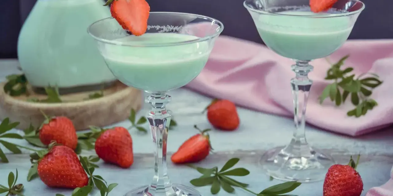 Zwei Gläser mit hellgrünem Likör, garniert mit Erdbeeren. Ein Krug mit dem Likör und frische Erdbeeren deuten darauf hin, dass man Waldmeister Schnaps selber machen kann.