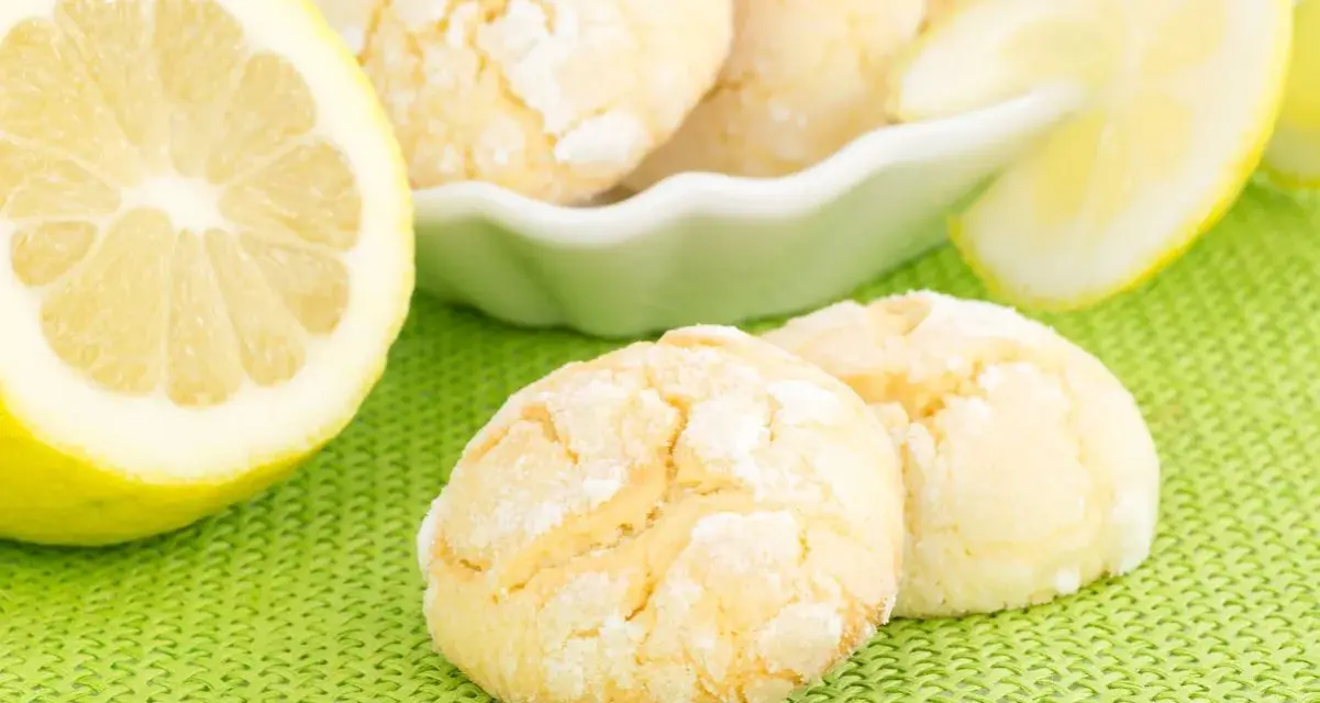 Biscotti al limone spolverati di zucchero a velo, accanto a fette di limone fresco su sfondo verde.