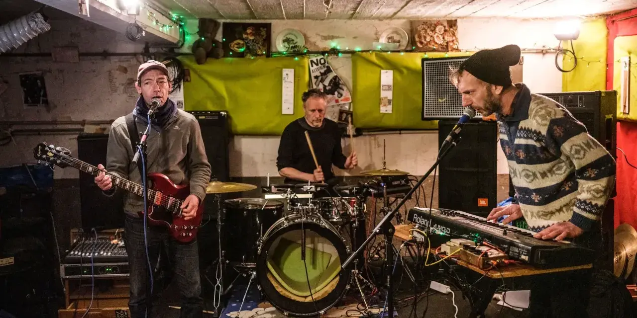 Un groupe de musique punk des années 90 joue dans un local sombre. Un guitariste, un batteur et un claviériste sont sur scène.