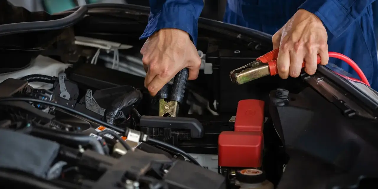 Hände mit Starthilfekabeln, um die **Batterie richtig anzuklemmen**. Ein Mechaniker in blauer Arbeitskleidung verbindet die Klemmen mit der Autobatterie.