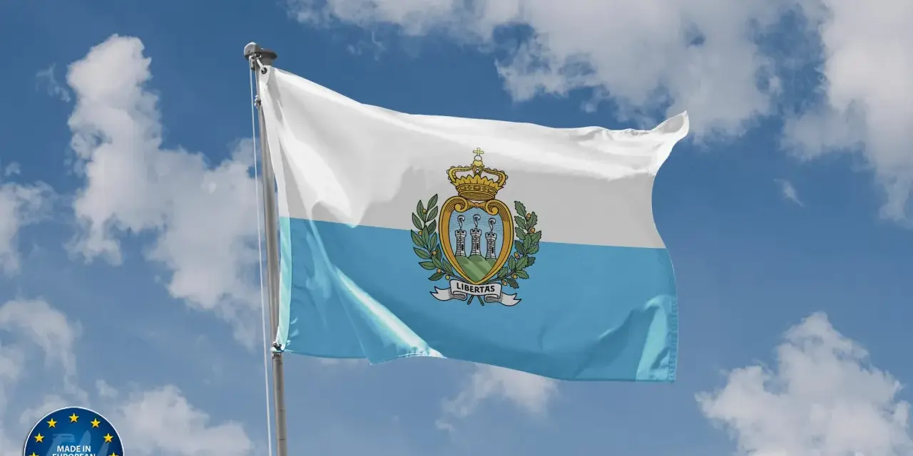Flaga San Marino powiewa na tle błękitnego nieba z chmurami. Herb z trzema wieżami i koroną zdobi biało-błękitny materiał.