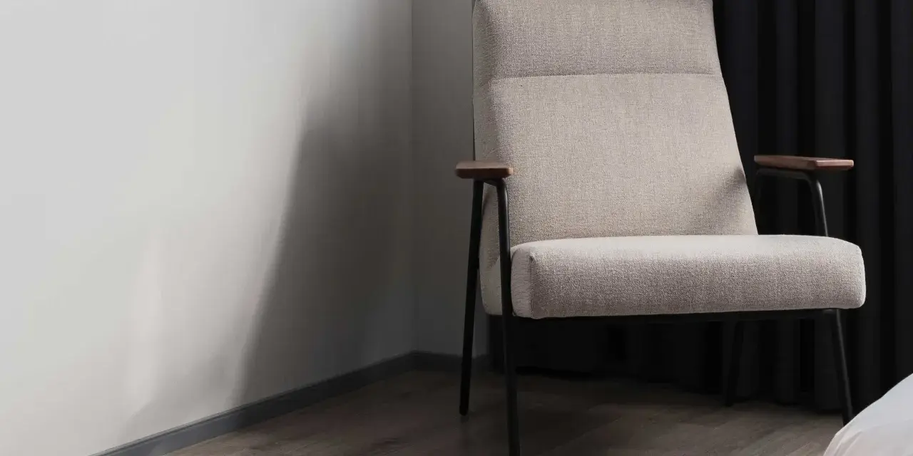 Fauteuil moderne avec un élégant tissu assise chaise, pieds fins et accoudoirs en bois. Il est placé près d'un mur blanc et de rideaux sombres.