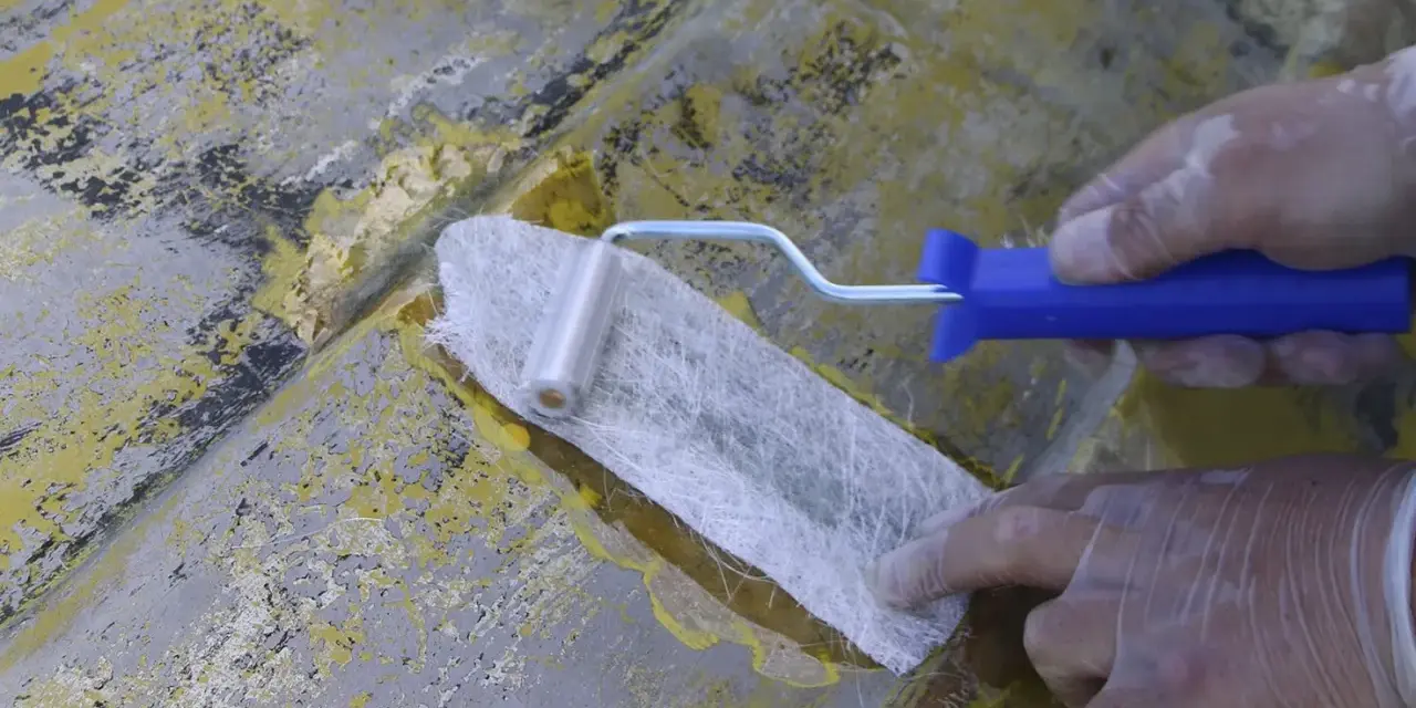 Händer med handskar arbetar med glasfibermatta och en roller för att laga en skadad yta.