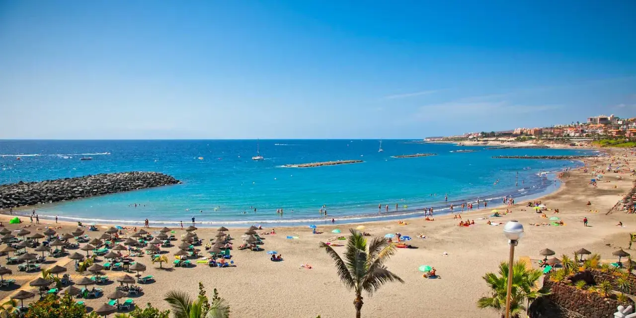 Spiaggia affollata con ombrelloni e lettini, mare azzurro e città costiera sullo sfondo. Sembra l'isola più bella delle Canarie.