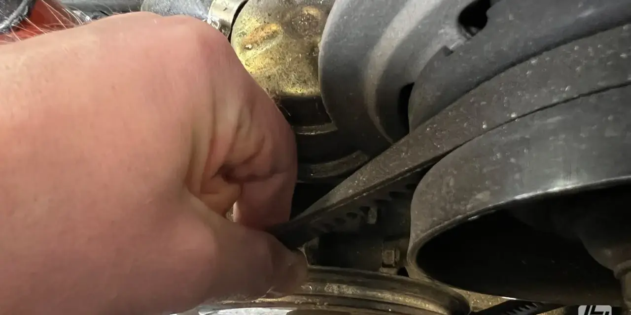 Hand prüft Keilriemen, der beim Anfahren quietscht. Der Riemen läuft über mehrere Riemenscheiben im Motorraum.