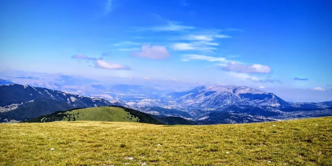 Prato verdeggiante con vista panoramica sul monte Pizzalto e le montagne circostanti sotto un cielo azzurro con nuvole sparse.