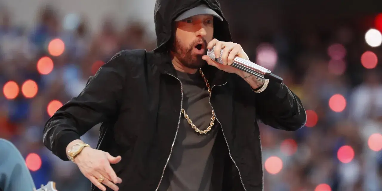 Eminem w czarnej bluzie z kapturem śpiewa do mikrofonu, w złotym łańcuchu i zegarku.
