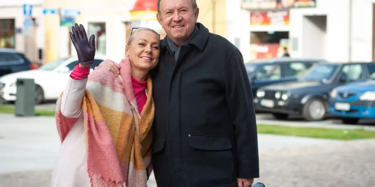 Małgorzata Szeptycka wita się z uśmiechem, obejmując partnera. W tle miejski krajobraz z samochodami.