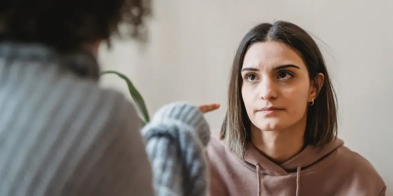 Dwie kobiety rozmawiają. Jedna wskazuje palcem na drugą, która patrzy z wyrzutem, jakby miała potworne wyrzuty sumienia.