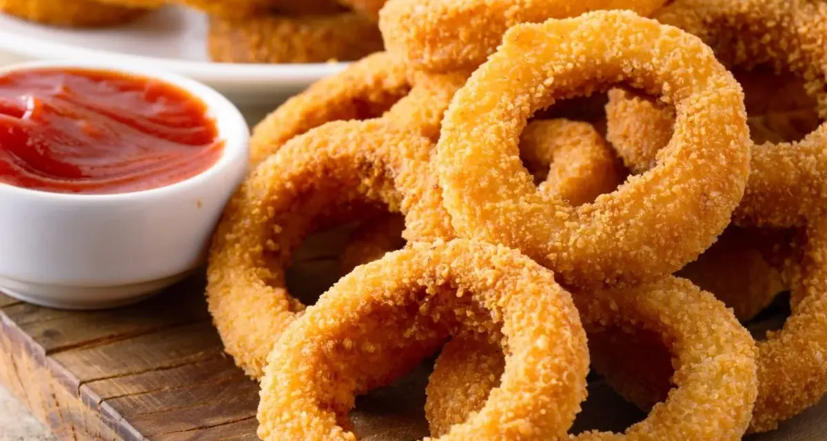 Aros de cebolla dorados y crujientes apilados junto a un tazón de salsa de tomate.