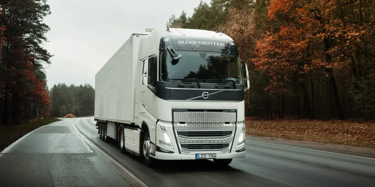 Biała ciężarówka Volvo FH12 z naczepą jedzie po mokrej drodze wśród jesiennego lasu.
