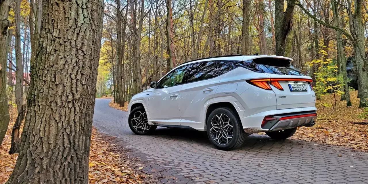 Biały Hyundai Tucson Hybrid na jesiennej drodze wśród drzew.