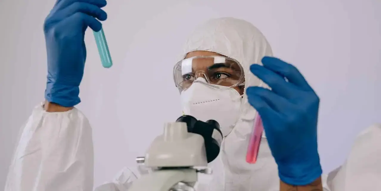 Badacz w kombinezonie i masce analizuje próbki w probówkach, przygotowując się do pracy w laboratorium chemicznym.