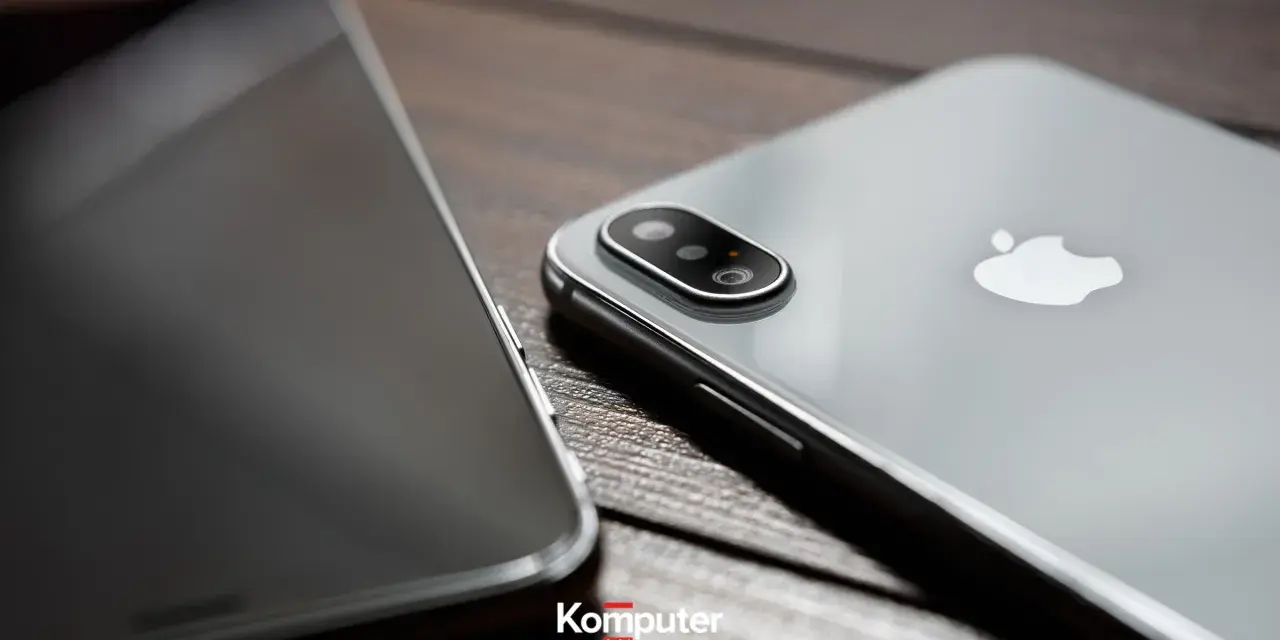 Dwa telefony leżą obok siebie na drewnianym blacie. Jeden z nich, srebrny iPhone, ma widoczny moduł aparatu. Drugi, z ciemnym ekranem, jest ustawiony bokiem. Idealne do rozmowy przez **Facetime**.