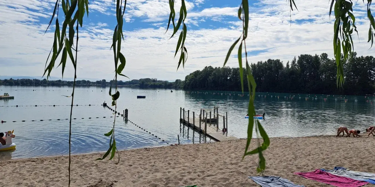 Ein sonniger Tag am Lauterbourg See. Handtücher liegen am Sandstrand, Kinder planschen im Wasser.