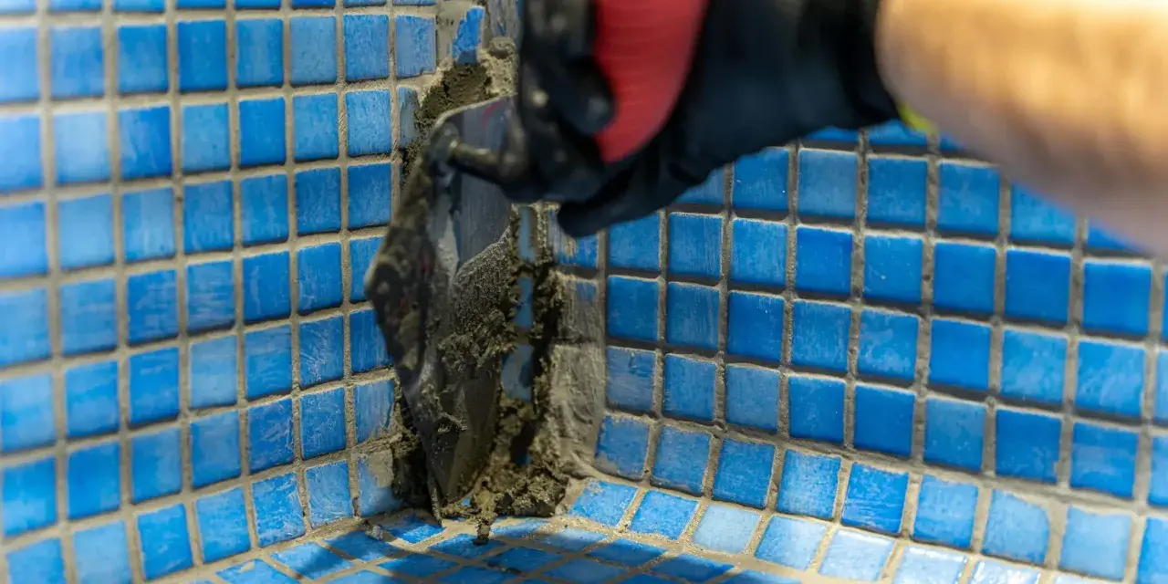 Application de mortier pour l'étanchéité d'une piscine en béton. Un ouvrier en gant noir et rouge applique le produit avec une spatule sur les carreaux bleus.