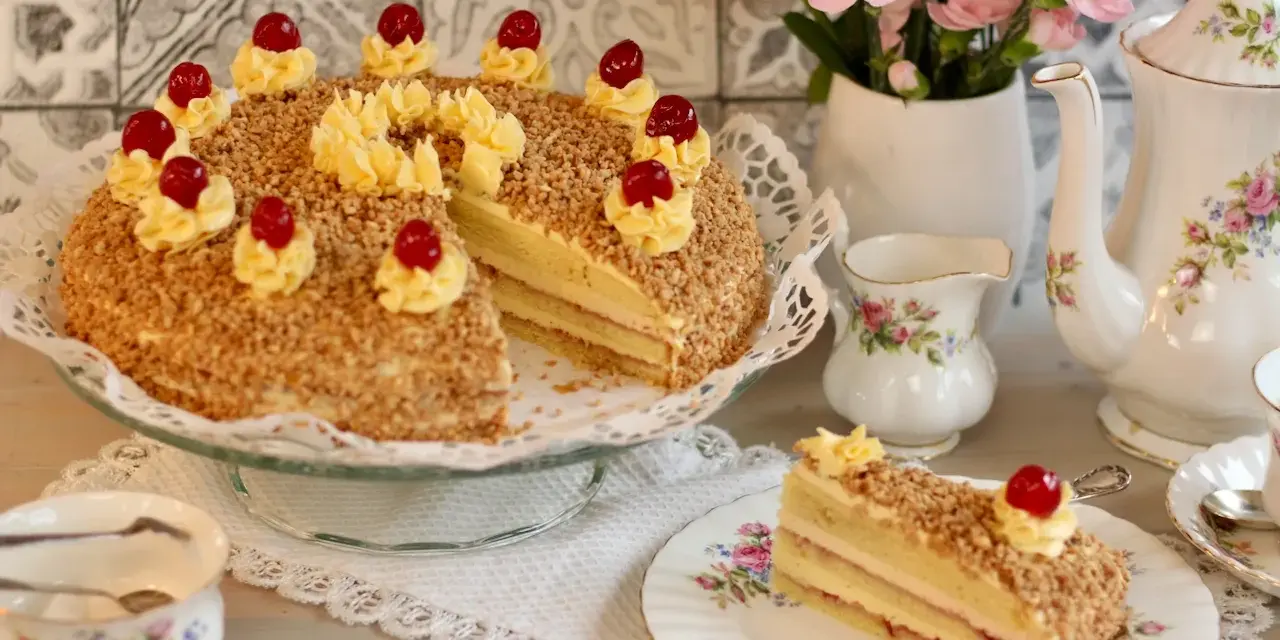 Ein köstlicher Frankfurter Kranz, verziert mit Buttercreme-Tupfern und Kirschen, umgeben von gehackten Nüssen. Ein Stück ist auf einem Teller angerichtet.