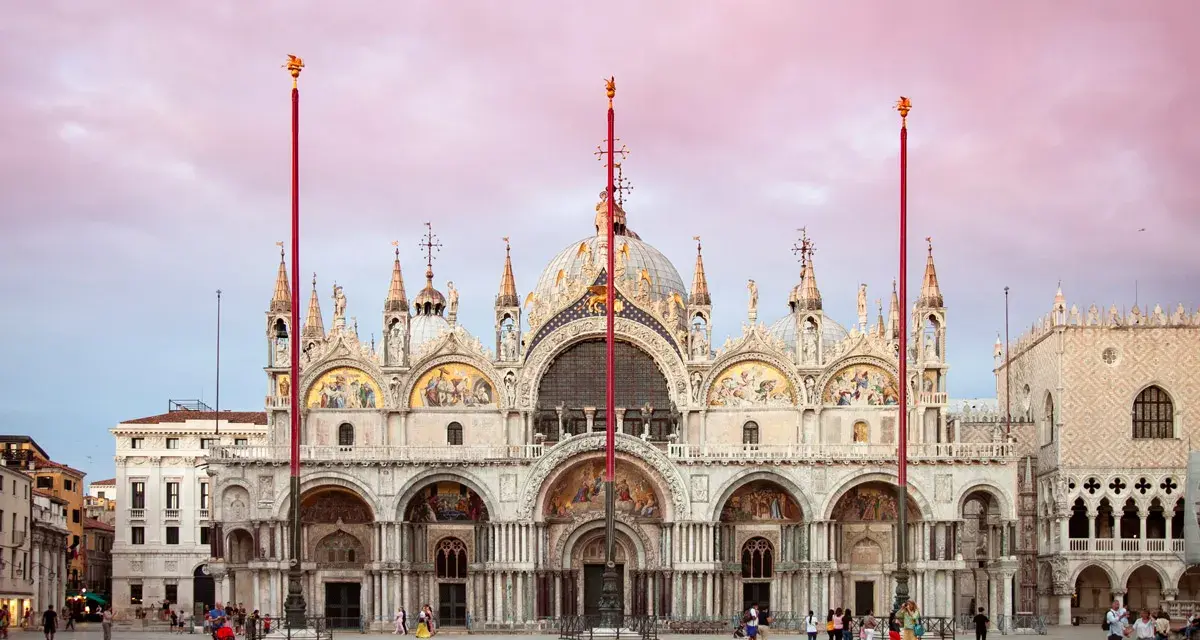 La maestosa basilica di San Marco a Venezia, con le sue cupole e mosaici dorati, si staglia sotto un cielo rosa al tramonto.