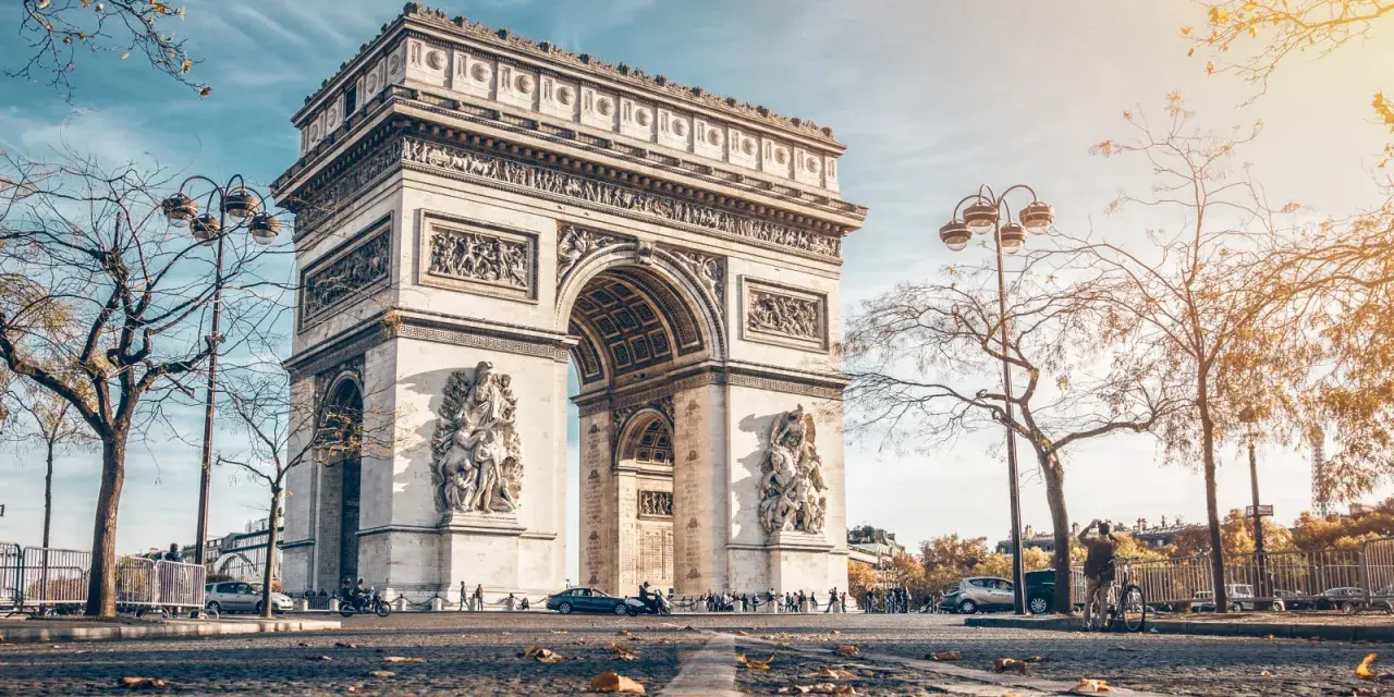 La majestuosa entrada del Arco del Triunfo en París, con esculturas y detalles ornamentales, bajo un cielo azul y árboles otoñales.
