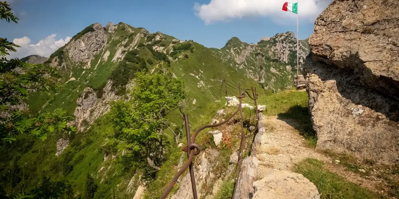 Sentiero della Pace tra montagne verdi, con bandiera italiana che sventola. Un percorso che ricorda la storia.