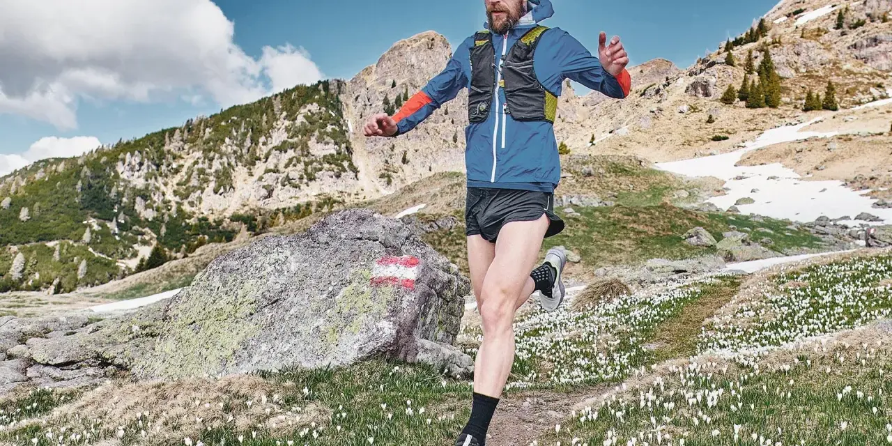 Uomo con barba corre in montagna, circondato da crochi bianchi e rocce. Il sentiero si snoda tra prati e pendii innevati.