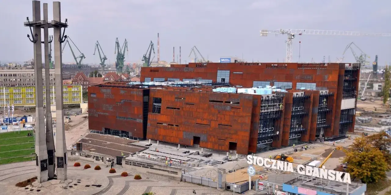 Nowoczesne muzeum na terenie Stoczni Gdańskiej, z charakterystycznymi, rdzawymi fasadami, w otoczeniu dźwigów stoczniowych i Pomnika Poległych Stoczniowców.