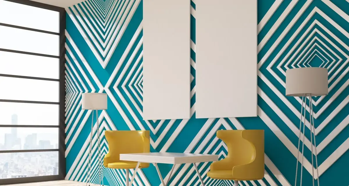 Modern room with striking **diseños pared geometrica** in teal and white. Two yellow chairs and a table sit before the wall, with floor lamps and blank canvases.